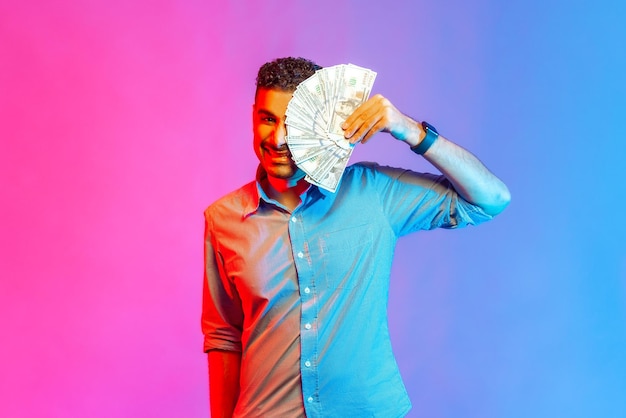 Portret bogatego zabawnego człowieka w koszuli zakrywającej połowę twarzy dolarami patrząc na kamerę z uśmiechem zębów, który jest szczęśliwy, aby wygrać na loterii Kryty studio strzał na białym tle na kolorowym tle światła neonowego