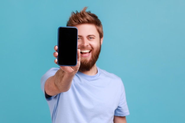 Portret brodatego mężczyzny trzymającego telefon komórkowy i patrzącego na kamerę z uśmiechem zębów i pozytywnym wyrazem zadowolenia z technologii Kryty studio strzał na białym tle na niebieskim tle
