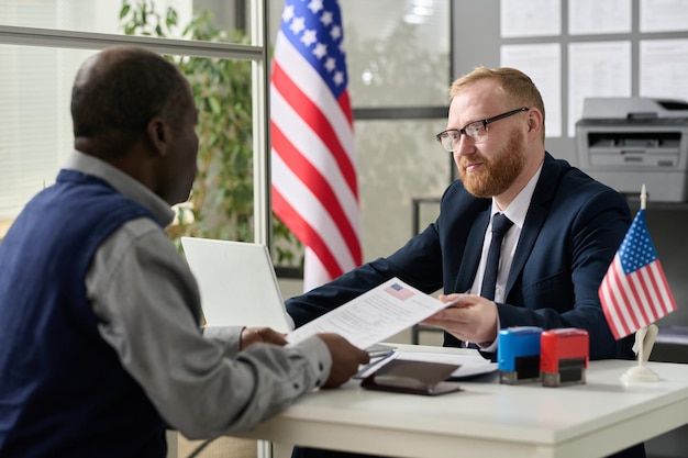 Portret brodatego pracownika płci męskiej wręczającego wniosek wizowy czarnemu mężczyźnie w amerykańskim biurze imigracyjnym