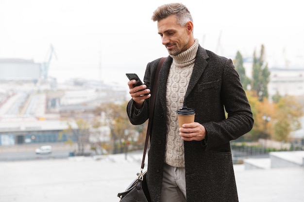 Portret brodaty mężczyzna 30s ubrany w kurtkę za pomocą telefonu komórkowego i trzymając kawę na wynos, idąc ulicą miasta