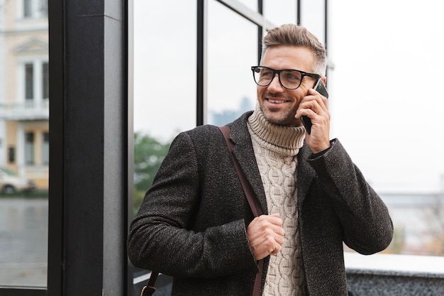 Portret brodaty mężczyzna 30s w okularach, spacerując ulicą miasta i przy użyciu smartfona