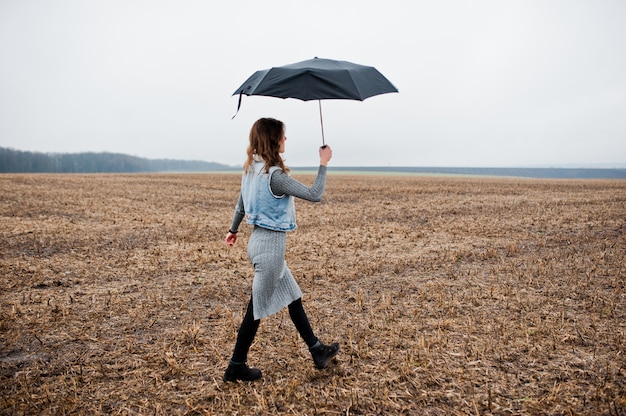 Portret brunetki kędzierzawa dziewczyna w cajg kurtce z czarnym parasolem przy polem.