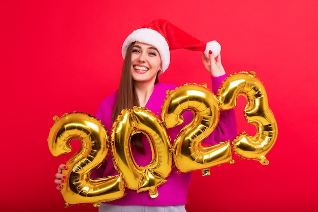 Portret brunetki w dobrym nastroju zawiera figury nowego roku 2023 Młoda kobieta założyła czapkę Świętego Mikołaja