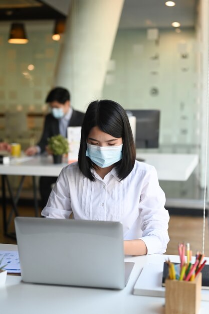 Portret businesswoman noszenie maski na twarz pracy z laptopem w biurze.