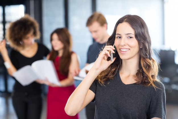 Portret Businesswoman Rozmawia Przez Telefon Komórkowy, Podczas Gdy Jej Koledzy Stoją Za Nią W Biurze