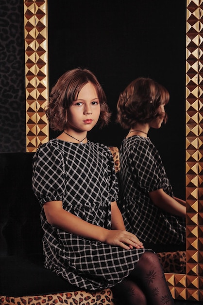 Portret całkiem ładna nastolatka w eleganckiej sukience przy lustrze w ciemnym stylowym wnętrzu salonu Dzieci emocje i pozowanie Koncepcja stylu moda i uroda Skopiuj miejsce na stronie