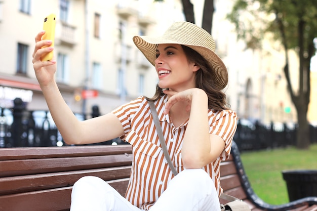 Portret całkiem młoda kobieta Dokonywanie selfie przez telefon, siedząc na ławce na ulicy miasta.