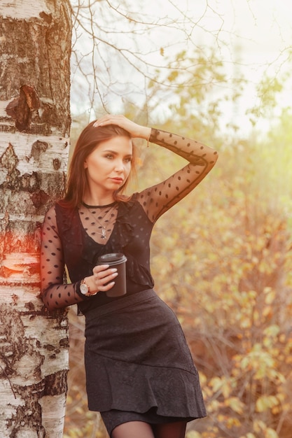 Zdjęcie portret całkiem młoda kobieta o słowiańskim wyglądzie w ciemnej sukni z filiżanką kawy jesienią, stojąca obok brzozy na tle jesiennego parku. złota jesień. skopiuj miejsce