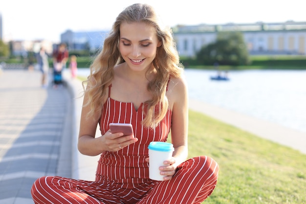 Portret całkiem młoda kobieta siedzi na brzegu rzeki z nogami skrzyżowanymi w letni dzień, przy użyciu smartfona.