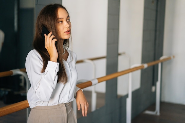 Portret całkiem młoda kobieta w stylowych ubraniach rozmawia przez telefon komórkowy, stojąc w pokoju z lustrem, odwracając wzrok Szczęśliwy atrakcyjny udany nauczycielka pozuje w klasie sztuki