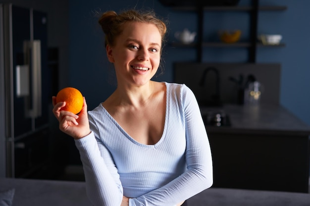 Portret całkiem roześmianej kaukaskiej dziewczyny trzymającej całą świeżą soczystą pomarańczę stojącą w domu w jej kuchni Młoda szczęśliwa kobieta trzyma pomarańczowy owoc stojący na stylowym tle nowoczesnej kuchni