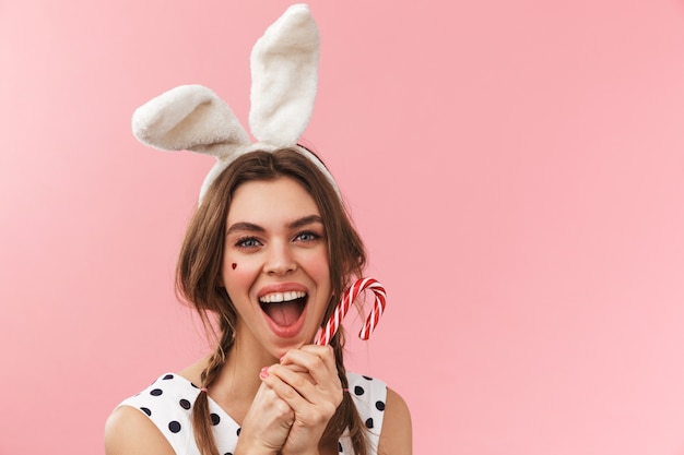 Portret całkiem uroczej dziewczyny noszącej uszy królika stojących na białym tle, krzywiąc się, trzymając candy cane