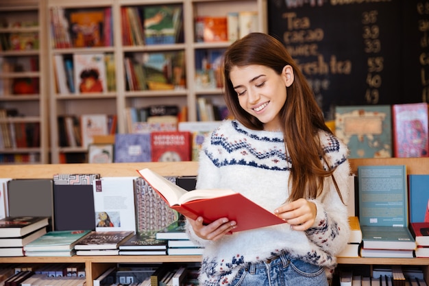 Portret całkiem uśmiechniętej dziewczyny czytającej książkę w pomieszczeniu w bibliotece