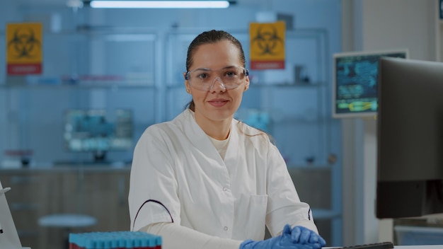 Portret chemika w okularach ochronnych uśmiecha się do kamery, gdy pracuje na komputerze, by odkryć lek w laboratorium. Mikrobiolog w okularach i pracujący nad eksperymentem naukowym.