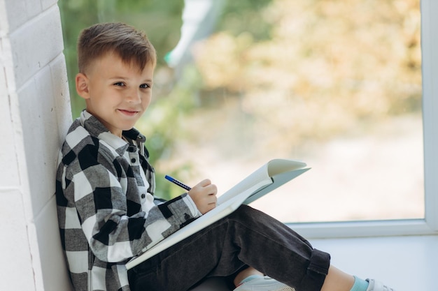 Portret chłopca odrabiającego pracę domową Chłopiec siedzi na parapecie i pisze w zeszycie Uczeń odrabia pracę domową przy oknie Nauka w domu