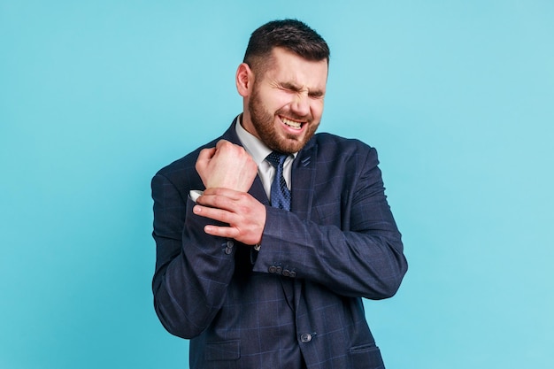 Portret chorego biznesmena w garniturze w oficjalnym stylu stojącym z grymasem bólu masującym ból nadgarstka, cierpiący na kontuzję ręki lub zwichnięcie. Kryty studio strzał na białym tle na niebieskim tle