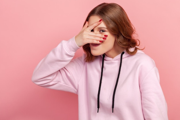 Portret ciekawa nastolatka kobieta w bluzie z kapturem szpiegostwo ukrywanie i podglądanie przez palce, szukając tajemnic plotek Kryty studio strzał na białym tle na różowym tle