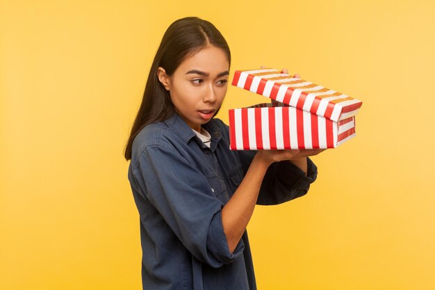 Portret ciekawej śmiesznej dziewczyny w denimowej koszuli otwierającej prezent i patrzącej z zainteresowaniem do pudełka, zaglądając do środka w oczekiwaniu na miłą niespodziankę. strzał w studio na białym tle na żółtym tle