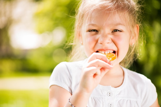 Portret cute little girl stojący w parku i trzymając w ręku pomarańczowy Lollipop.