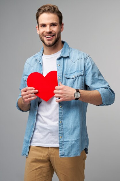 Portret czarującego mężczyzny trzymaj rękę papierową kartę w kształcie serca nosić koszulę w stylu casual na białym tle na szarej ścianie
