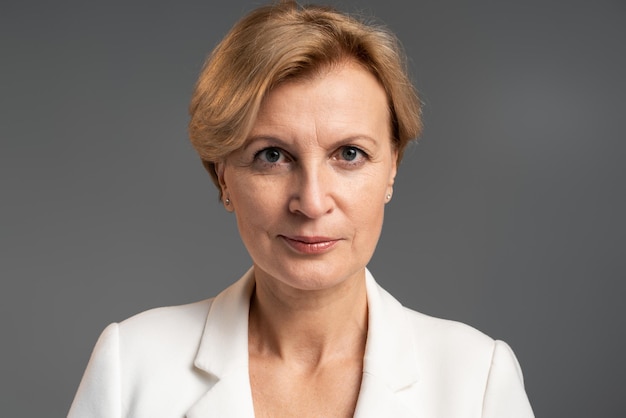 Portret dobrze ubranej dojrzałej atrakcyjnej bizneswoman patrzącej w kamerę ze spokojnym poważnym wyrazem twarzy podczas pozowania nad szarą ścianą