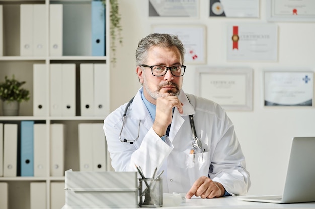 Portret dojrzałego lekarza w białym fartuchu, patrzącego na kamerę, siedząc w swoim miejscu pracy z laptopem
