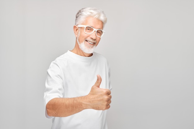 Portret dojrzałego mężczyzny w białej koszulce i okularach uśmiechającego się radośnie pokazującego kciuk w górę gestem izolowanym na białym tle studia Zatwierdza dobry wybór, właściwą decyzję