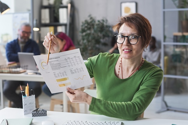 Portret dojrzałej bizneswoman w okularach, wskazując na dokument i pokazujący wyniki biznesowe, siedząc w swoim miejscu pracy