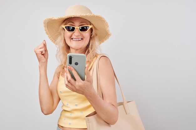 Portret dojrzałej blondynki w słomkowym kapeluszu, żółtym topie i okularach przeciwsłonecznych z smartfonem i torbą na zakupy na białym tle studia