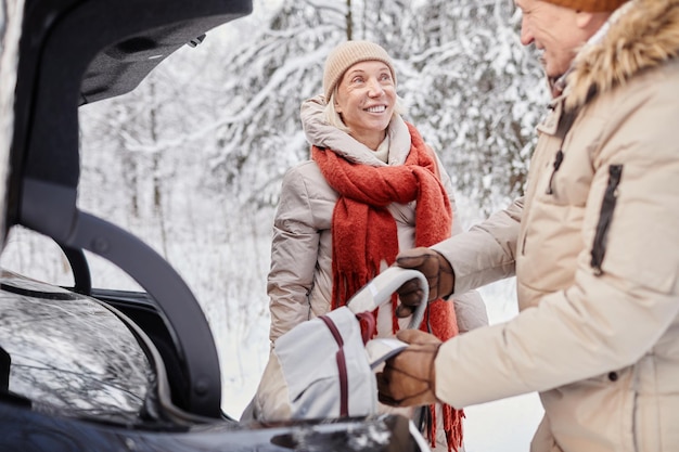 Portret dojrzałej pary cieszącej się zimowym wypadem na łonie natury, stojąc samochodem z otwartym bagażnikiem