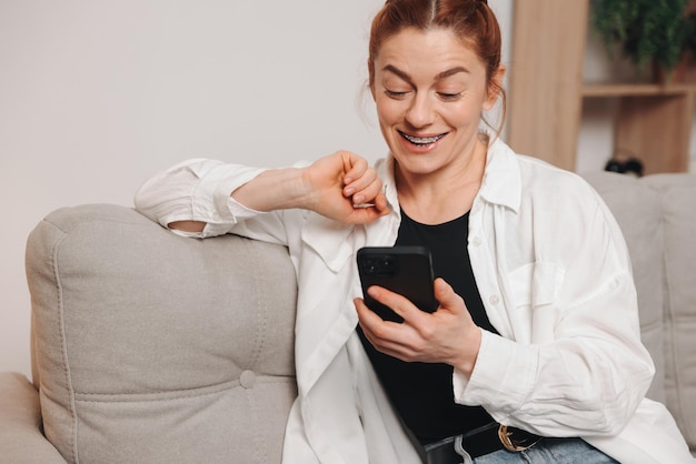Portret dojrzałej uśmiechniętej kobiety z aparatem ortopedycznym używającej smartfona w domu