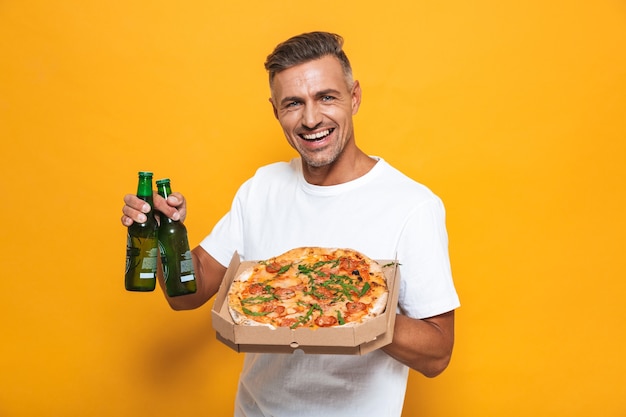 Portret dorosłego mężczyzny w wieku 30 lat w białej koszulce, pijącego piwo i jedzącego pizzę, stojąc odizolowane na żółto
