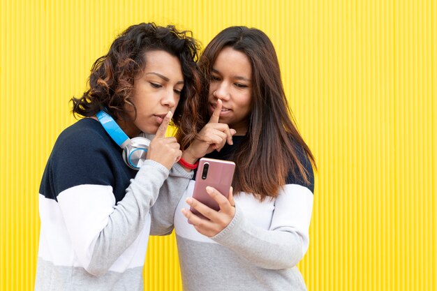 Portret dwóch brązowych dziewczyn na żółtym tle. Oboje wykonują gest prosząc o ciszę, patrząc na inteligentny telefon. Miejsce na tekst.