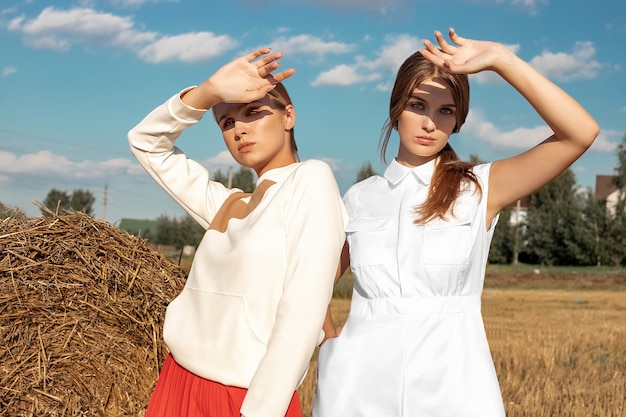 Portret dwóch jasnowłosych dziewcząt w modnych i stylowych ubraniach na tle pola i stosu słomy