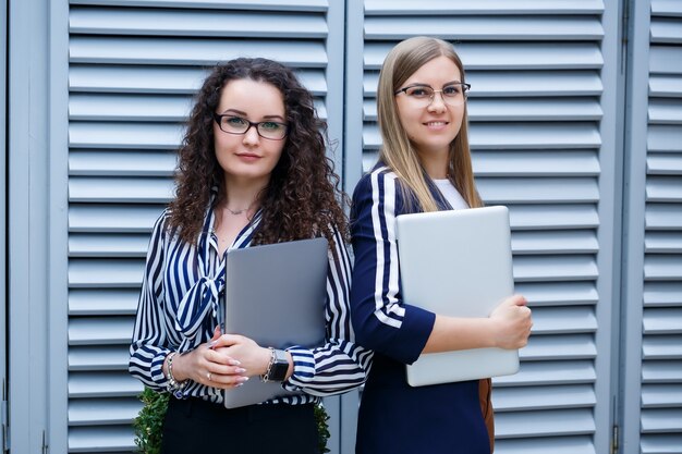 Zdjęcie portret dwóch kobiet biznesmenów, którzy stoją z laptopami w rękach, nowy projekt. młodzi biznesowi koledzy omawiają pracę biurową. pomyślny menedżer żeński w okularach. pomysł na biznes