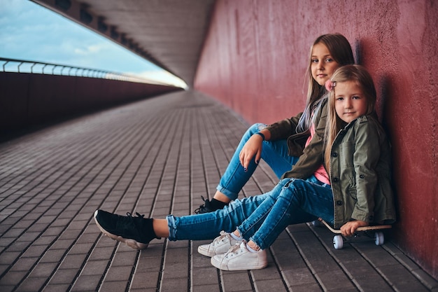 Portret dwóch małych sióstr ubranych w modne ubrania, opierając się o ścianę, siedząc na deskorolce na chodniku mostu.