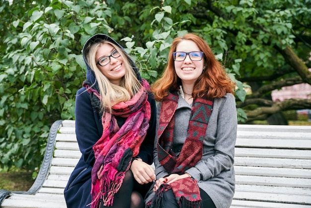 Portret Dwóch Młodych Atrakcyjnych Dziewczyn Rasy Kaukaskiej, Ubranych W Płaszcz, Siedzących Na ławce W Parku Miejskim Na Tle Zielonych Liści.
