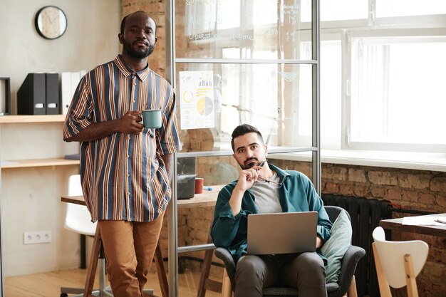 Portret dwóch młodych biznesmenów z laptopem i filiżanką kawy patrzących na kamerę podczas pracy w biurze