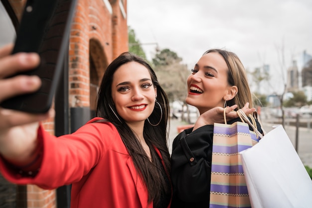 Portret dwóch młodych przyjaciół korzystających ze wspólnych zakupów podczas robienia selfie z telefonem na ulicy.