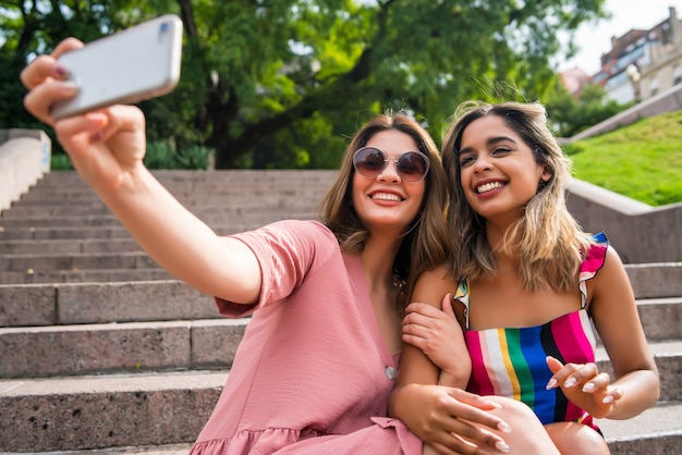 Portret dwóch młodych przyjaciół, uśmiechając się i biorąc selfie z telefonem komórkowym, siedząc na zewnątrz. Koncepcja miejska.