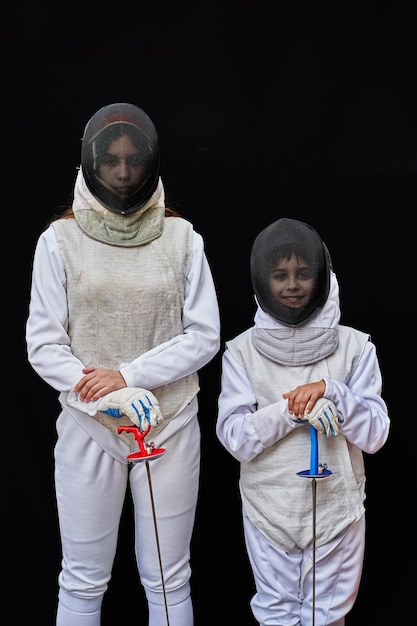 Portret dwóch młodych szermierzy ubranych w biały kostium szermierza i maskę, trzymających przed sobą miecz. Na białym tle na czarnym tle