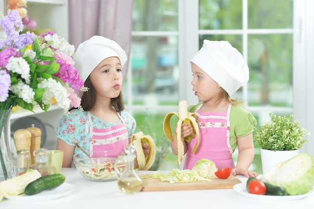 Portret dwóch uroczych dziewczynek jedzących banany w kuchni w fartuchach