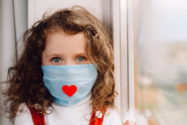Portret dziecka w wieku przedszkolnym siedzi na parapecie w domu, ubrany w maskę wirusa z czerwonym sercem, mała dziewczynka patrzy na aparat. Epidemiczny pandemiczny rozprzestrzeniający się koronawirus 2019-ncov. Koncepcja dzień pielęgniarki.