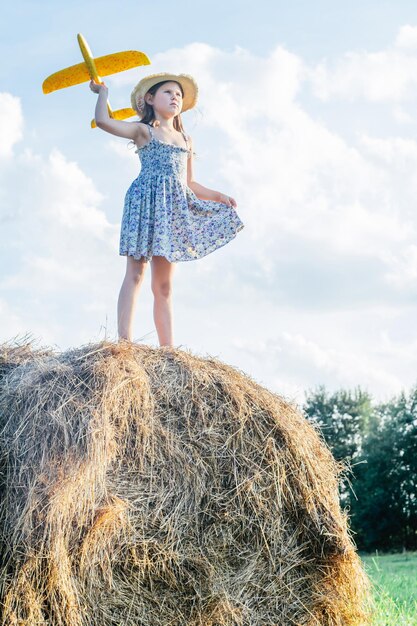 Portret dziewczynki, grając, stojąc na stogu siana w polu. Latanie, rzucanie żółtym samolocikiem. Jasny słoneczny dzień. Celowo koncepcja. Nowy początek, patrzenie w przyszłość, podróże. Mały kąt