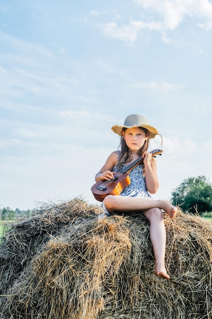 Portret dziewczynki grającej na małej gitarze ukulele, siedzącej na stogu siana na polu w pobliżu drzew, ubrana w sukienkę i kapelusz