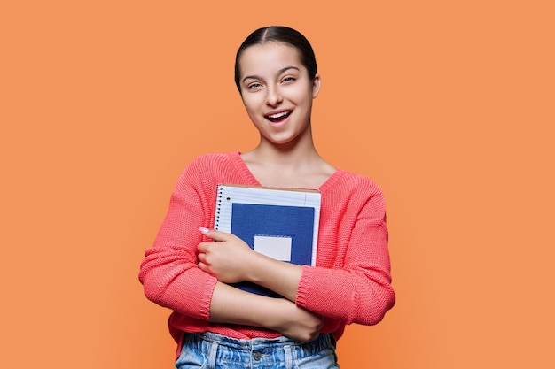 Zdjęcie portret dziewczynki, nastoletniej uczennicy szkoły średniej, na pomarańczowym tle studia uśmiechnięta uczennica 15-16 lat z książkami, zeszytami, patrząc na kamerę koncepcja wiedzy o edukacji w okresie dojrzewania