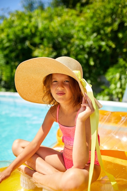 Portret dziewczynki w kapeluszu relaksuje się w basenie, pływa na nadmuchiwanym żółtym materacu