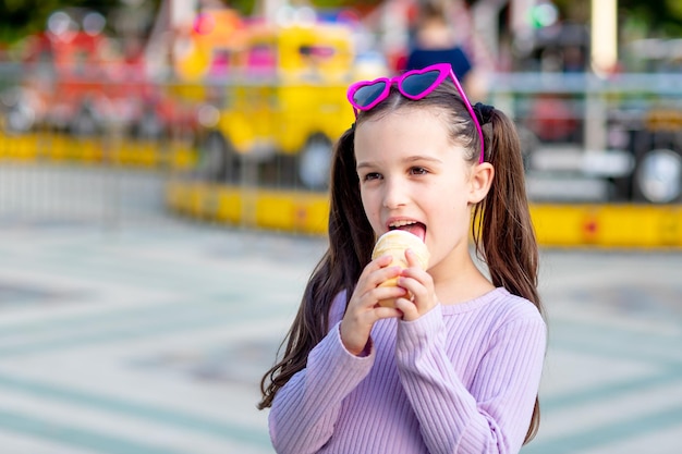 Portret dziewczynki w parku rozrywki latem jedzącej lody w pobliżu karuzeli i uśmiechającej się ze szczęścia w okularach przeciwsłonecznych koncepcja weekendów i wakacji szkolnych