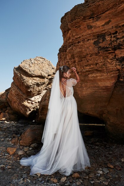 Portret dziewczynki w sukni ślubnej wspaniały pozowanie fotograf na plaży. Panna młoda jest na skałach