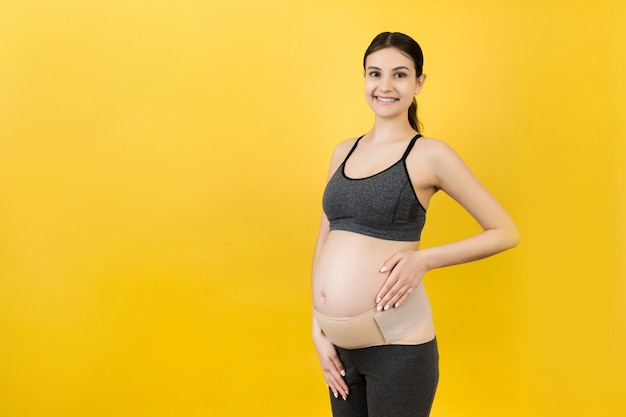 Portret elastycznej opaski macierzyńskiej ubranej na kobietę w ciąży, aby zmniejszyć ból pleców na żółtym tle z miejsca na kopię. Koncepcja ortopedycznego pasa podtrzymującego brzuch.